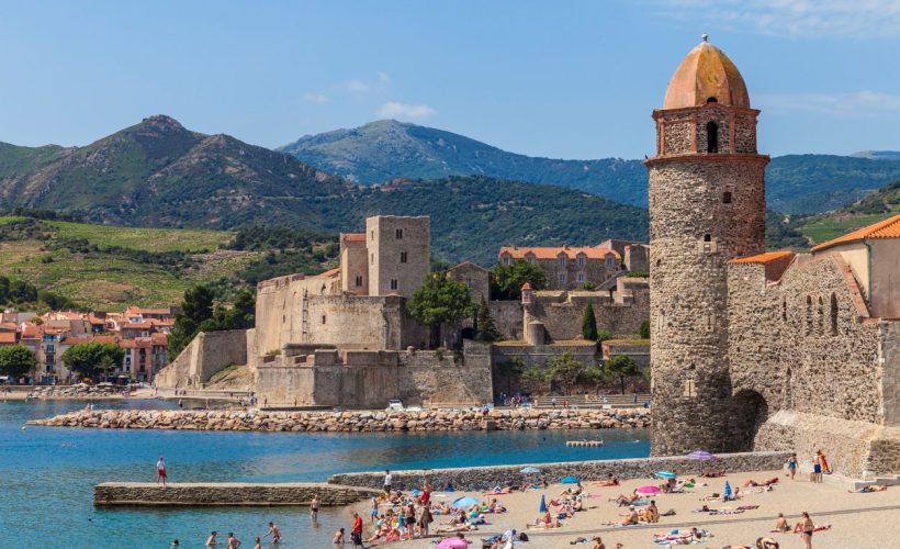 Collioure, France - July 01, 2016. View of the picturesque Collioure, situated in the Département Pyrénées-Orientales. Château Royal in the centre, lighthouse/spire on the right side. The beach is highly frequented by tourists and natives. Pyrenees Mountain Range in the background.